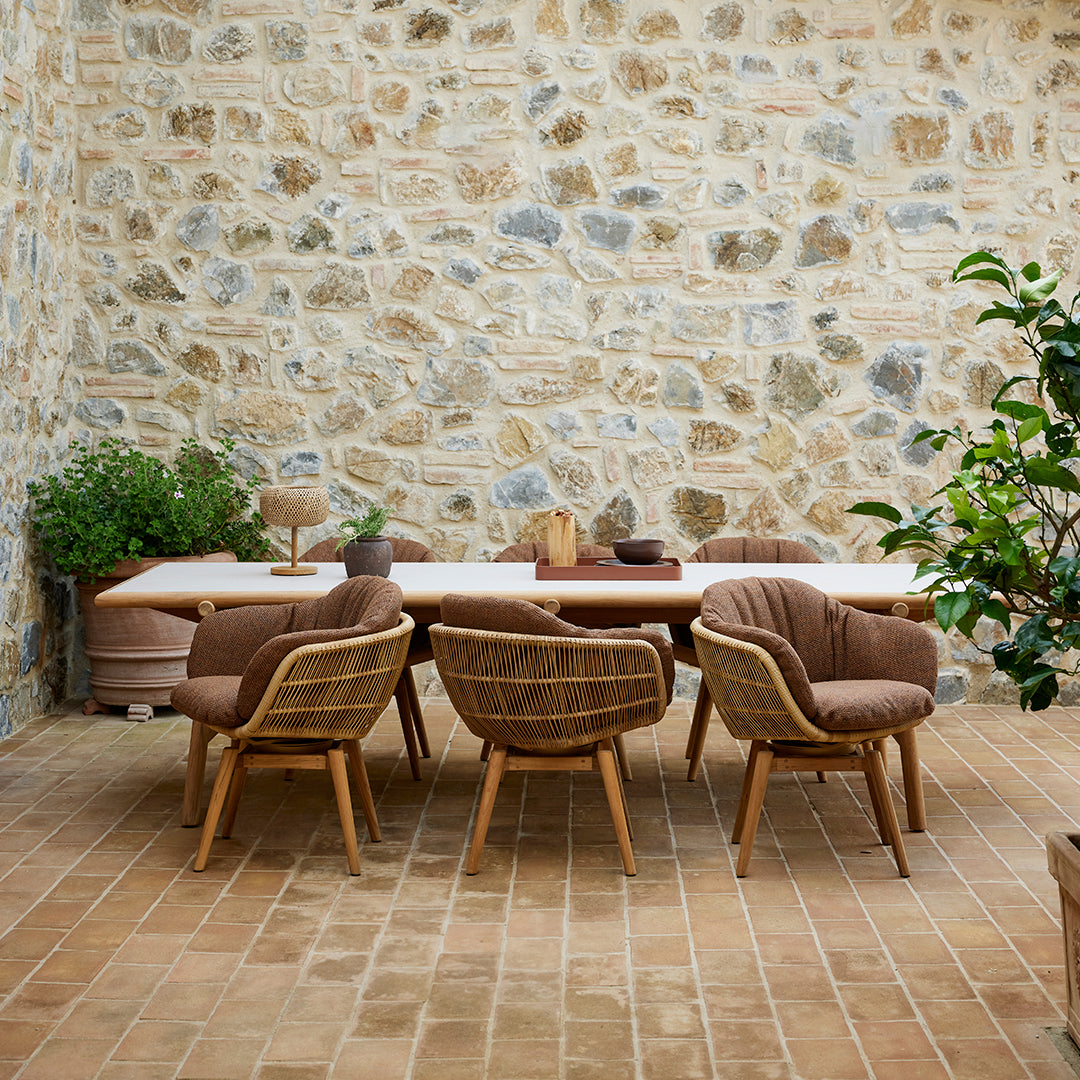 Natural wood dining table with beige and brown upholstered chairs against a stone wall and terracotta floor.