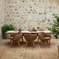 Natural wood dining table with beige and brown upholstered chairs against a stone wall and terracotta floor.