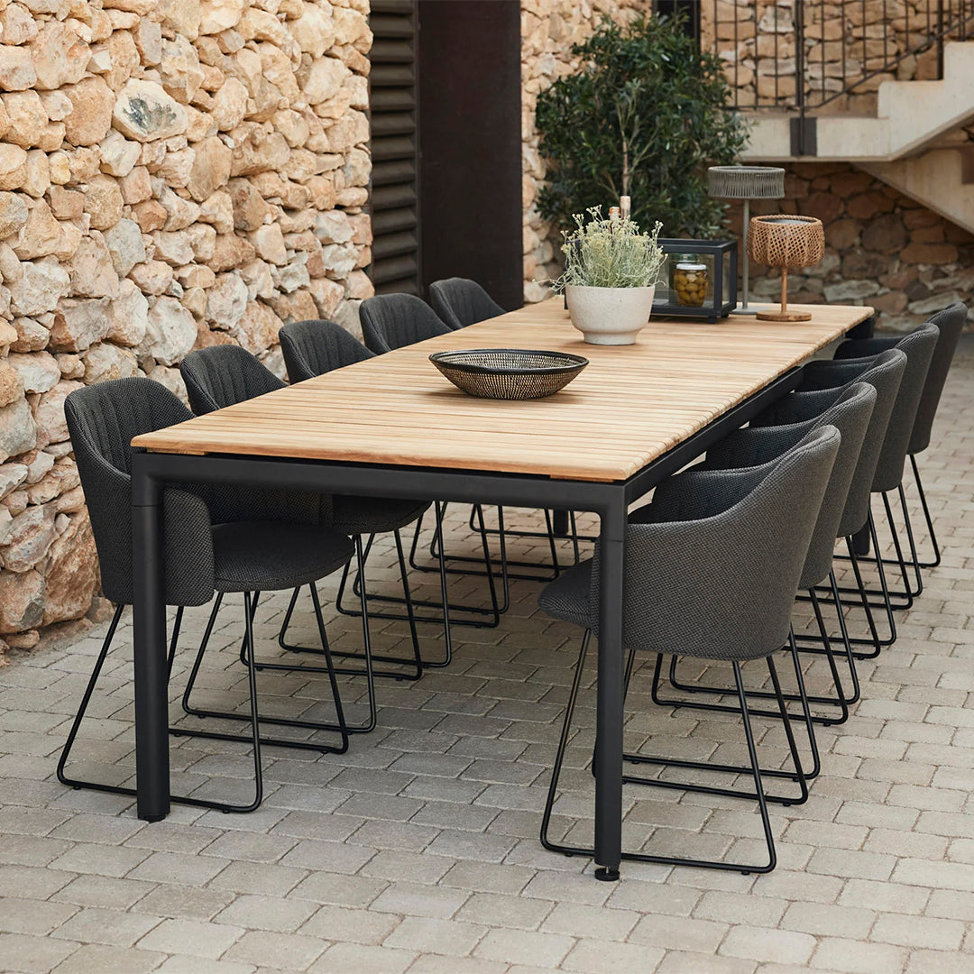Modern outdoor dining table with a natural wood surface, paired with sleek black metal legs and charcoal gray upholstered chairs.