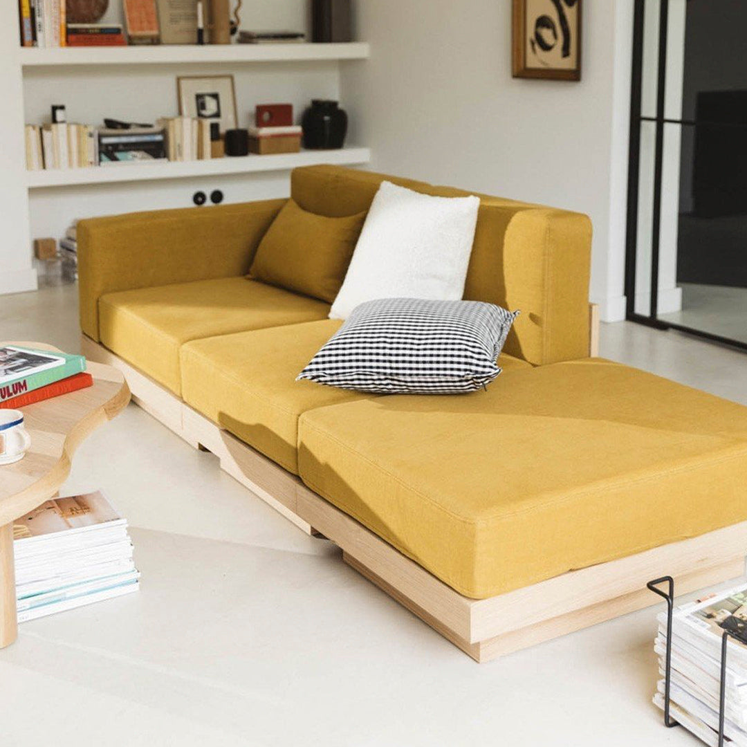 "Mustard yellow sectional sofa with a white and black checkered pillow on a minimalist wood base, set in a modern living space."