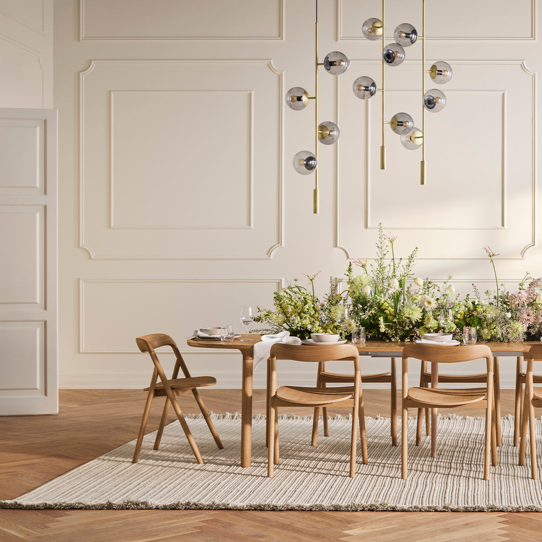 Light wood dining table with natural woven rug, surrounded by beige chairs and decorated with lush greenery and floral accents.