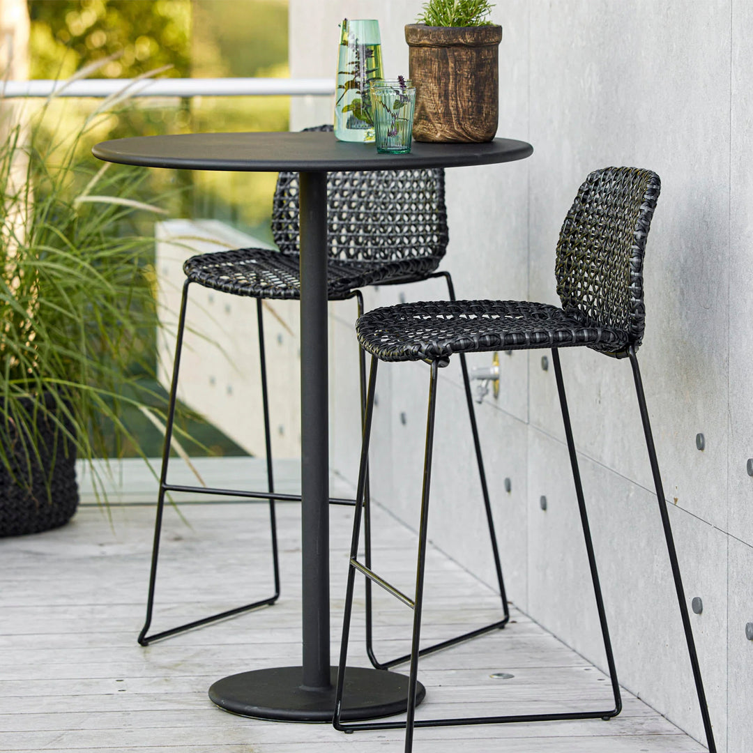 Black woven bar stools and a round table on a light wood deck with green plants and glass accents.