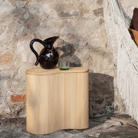 Natural wood finish side table with ribbed texture, paired with a sleek black pitcher and a glass. Earthy tones dominate.