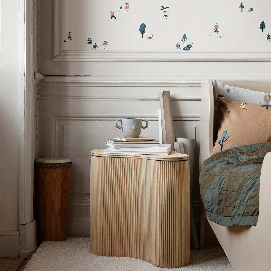 "Textured light wood nightstand with green quilt and playful patterned decor against soft, neutral wall."