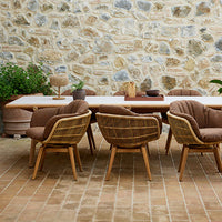 Brown wicker chairs with beige cushions surround a wooden table against a stone wall backdrop. Warm earthy tones.