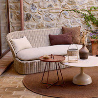 "Curved beige couch with brown cushions, round cream table, and textured brown rug against a stone wall backdrop."