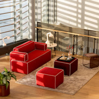 Red modern sofa and ottoman with white piping, set on a beige rug in a sunlit, contemporary living space.