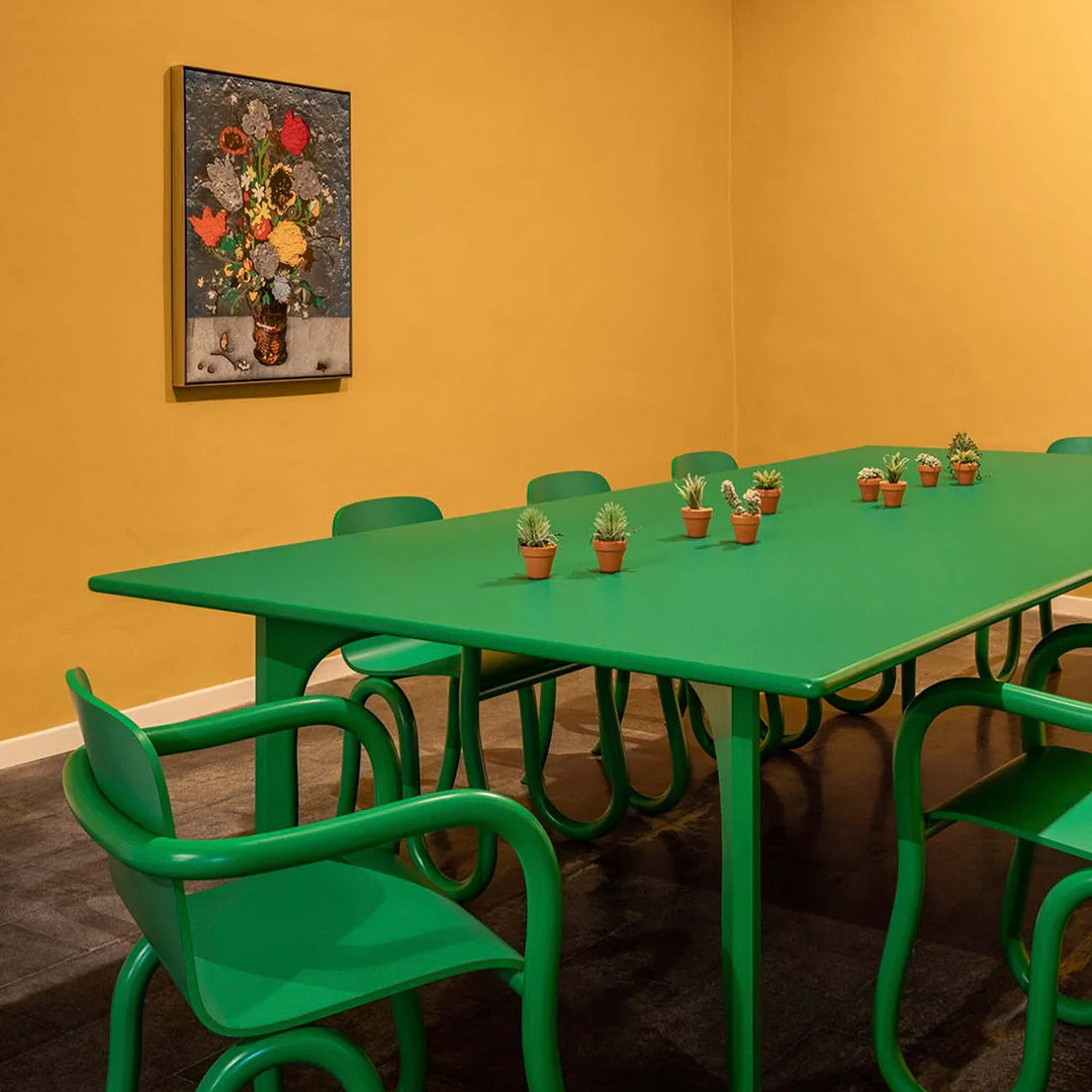 Green dining table and chairs with small potted plants on a vibrant yellow wall background. Modern, colorful design.