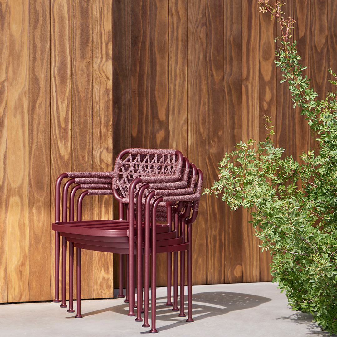 Stacked burgundy outdoor chairs with a lattice design against a warm wooden backdrop and green foliage.