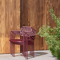 Stacked burgundy outdoor chairs with a lattice design against a warm wooden backdrop and green foliage.