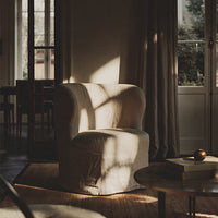 Curved beige armchair with soft texture, illuminated by natural light, casting gentle shadows on a wooden floor.