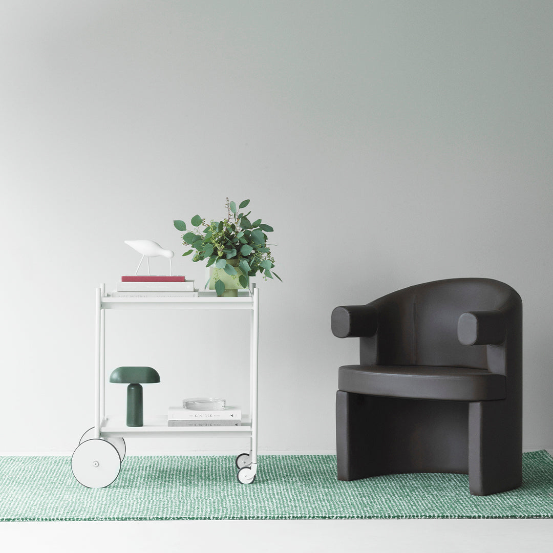 Black chair on green rug with white cart, featuring a simple pattern and lush green plant centerpiece.