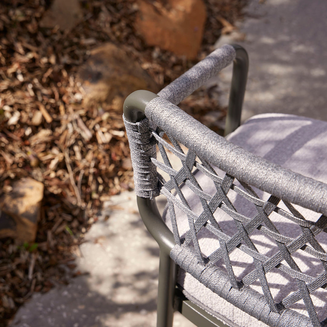 Gray woven chair with a subtle pattern, featuring a sleek dark frame and a soft cushion against natural stone backdrop.