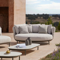 Light gray round outdoor sofa with plush cushions and a minimalist design, set against a natural landscape.