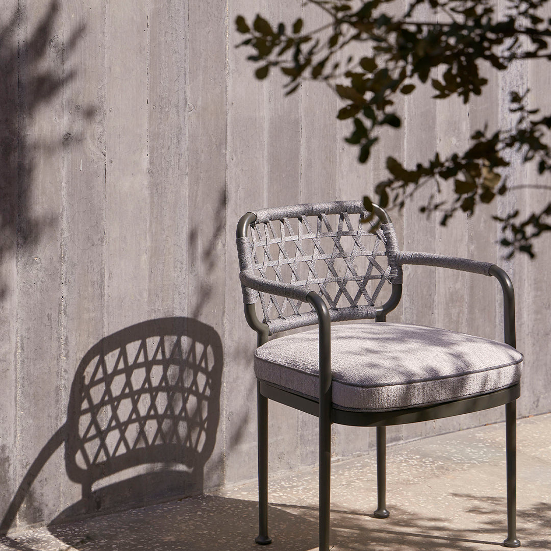 Gray outdoor chair with intricate woven pattern and soft beige cushion, casting a shadow on a textured surface.