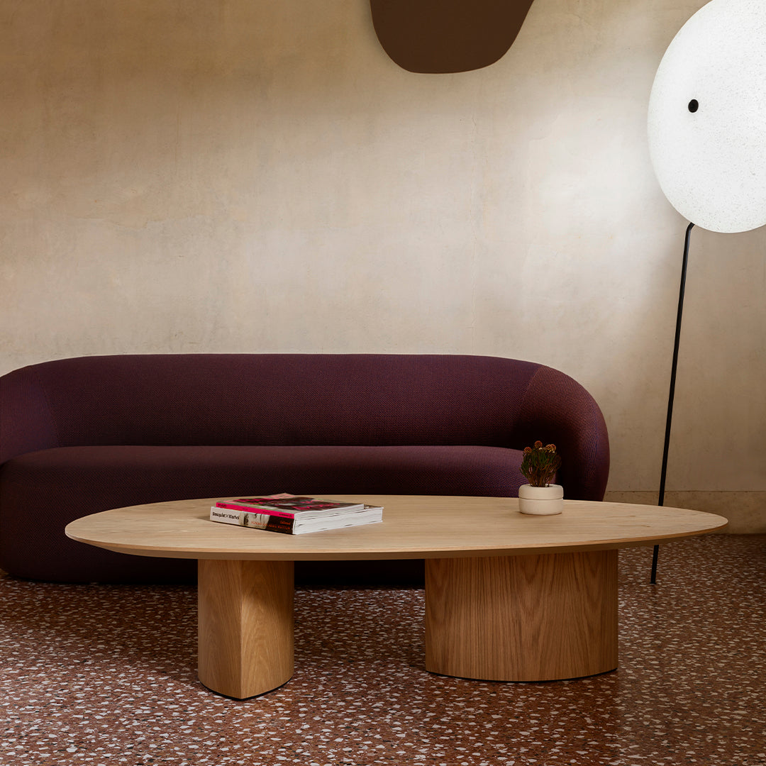 Purple curved sofa, natural wood oval table, and speckled clay floor create a cozy, modern living space ambiance.