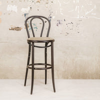 Black metal bar stool with a simple round wooden seat, featuring a classic bentwood design against a textured wall.
