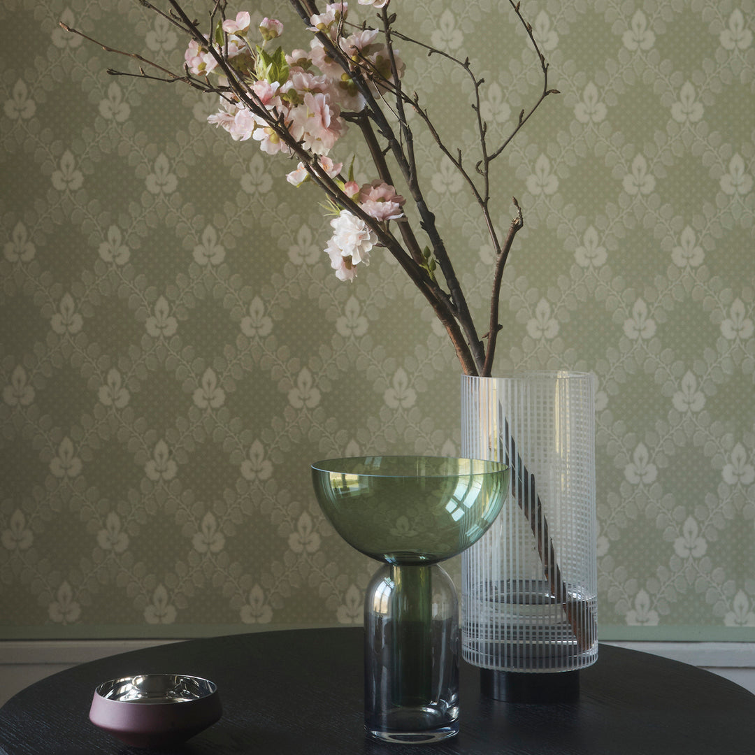 Green and transparent glass vases with pink cherry blossoms against a textured light green patterned wallpaper.