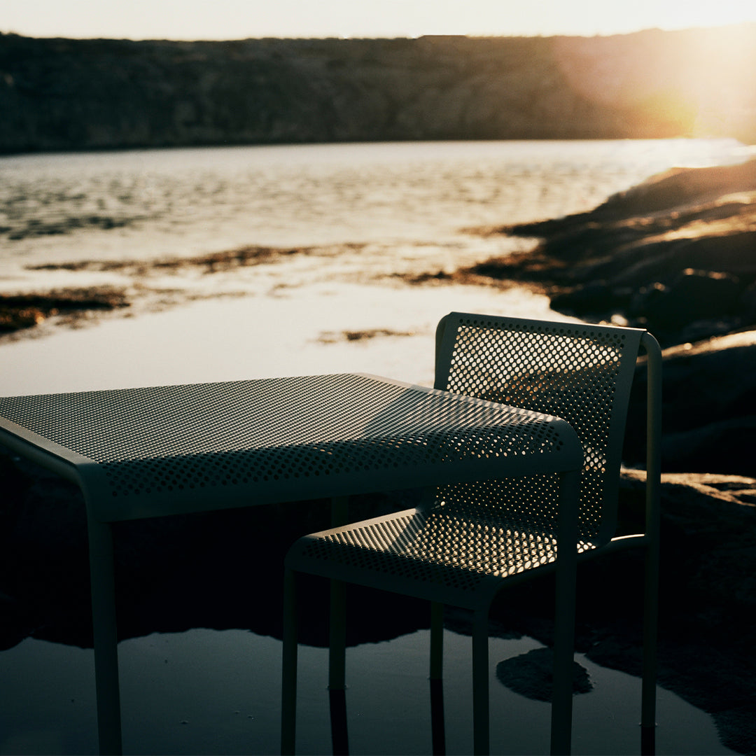 Light green perforated metal table and chair set near water at sunset, featuring a soft, captivating glow.