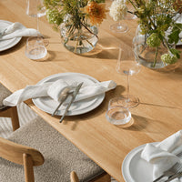 Elegant white dinnerware on a light wood table, adorned with colorful floral arrangements in clear vases.