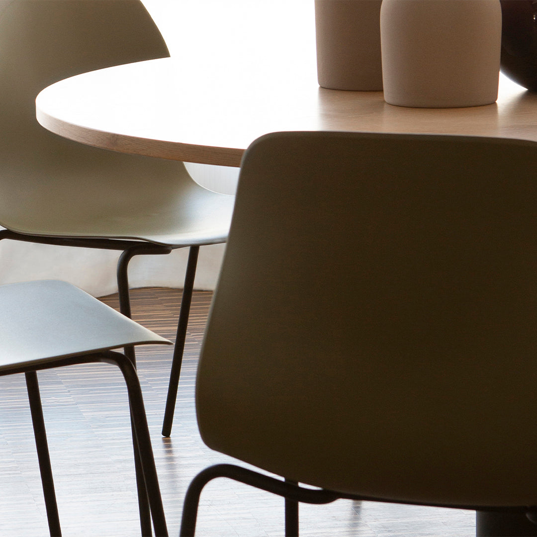 Minimalist dining set featuring soft olive-green chairs, a light wood table, and neutral-toned vases against a warm backdrop.