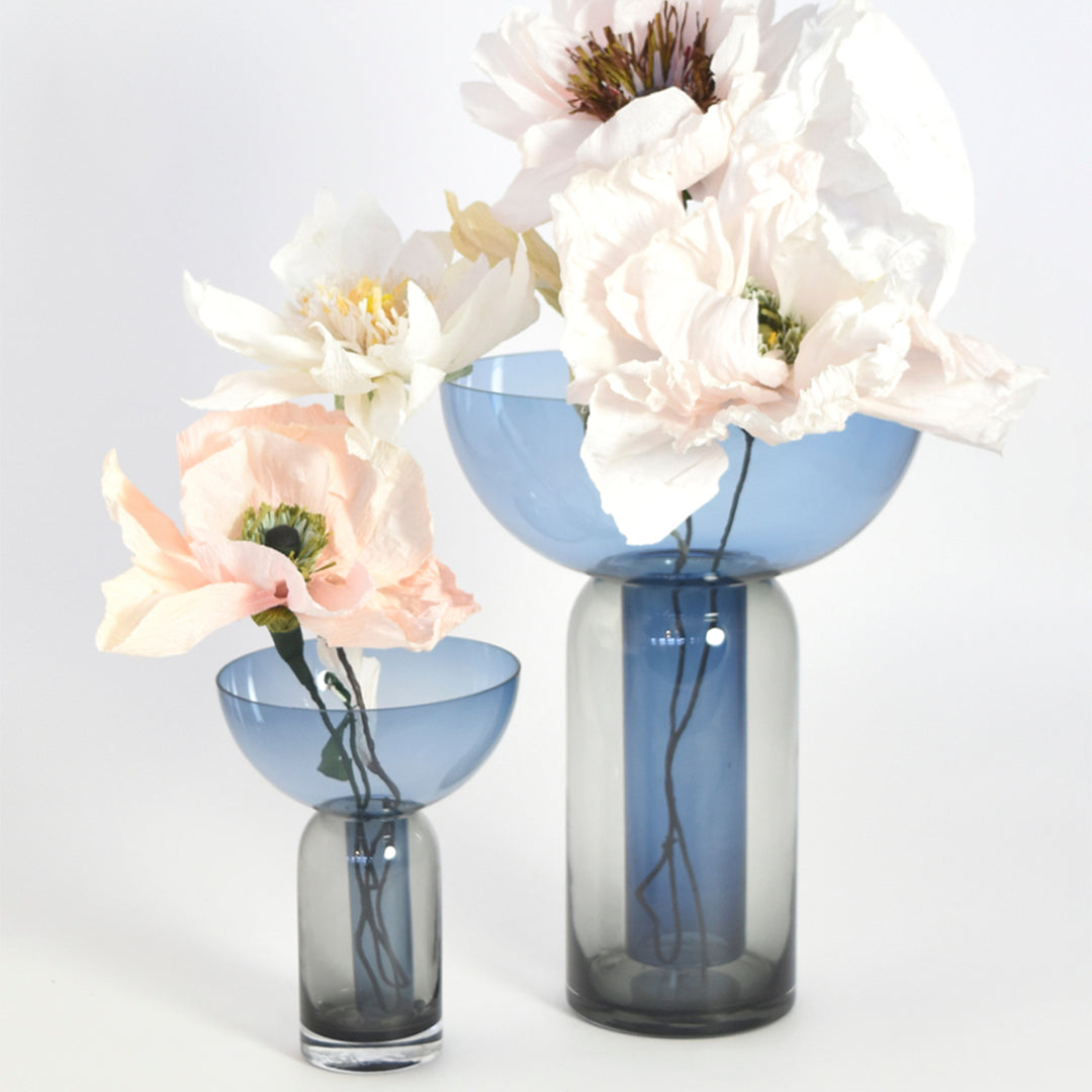"Blue and grey glass vases with delicate pastel flowers in soft pink and white, featuring elegant shapes and smooth finishes."