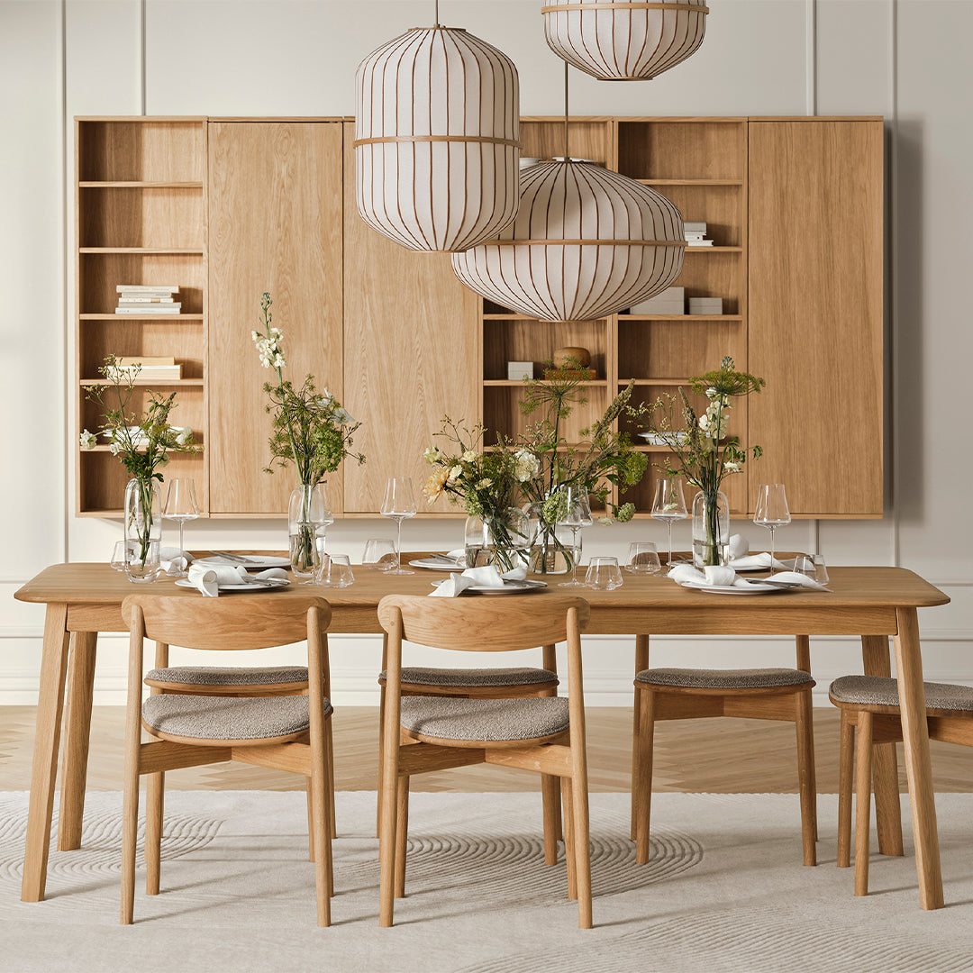 Wooden dining table with light oak finish, white and beige patterned lampshades, and neutral floral arrangements.