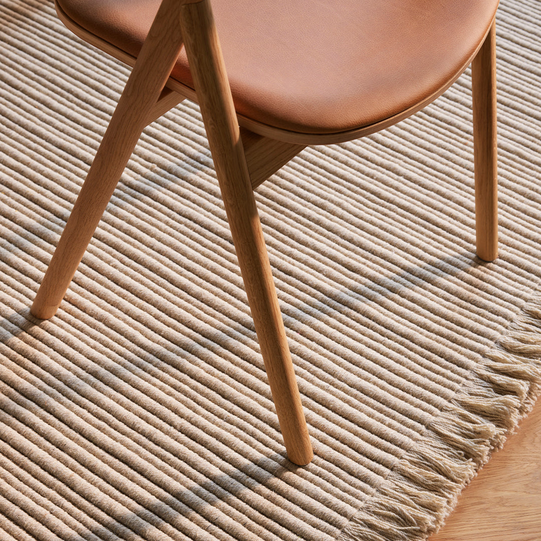 Natural beige woven rug with fringe detail, paired with a light tan chair, creating a warm, inviting aesthetic.
