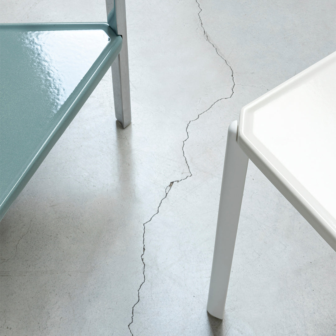 Two modern tables in soft teal and sleek white, showcasing glossy surfaces against a cracked concrete floor.