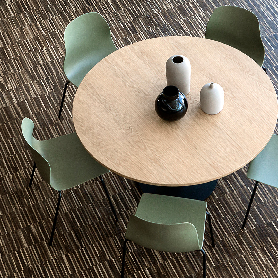Light wood round table with sage green chairs and black and white vases on a textured brown patterned floor.
