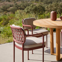 Rust-colored woven chairs with cream cushions, paired with a natural wood table against a green outdoor backdrop.
