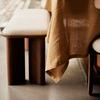 Light beige upholstered bench with dark wood legs, complemented by a warm, earthy-toned table linen draping.