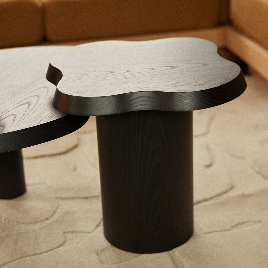 Black wooden coffee table with a unique scalloped edge and cylindrical base, set against a beige textured rug.