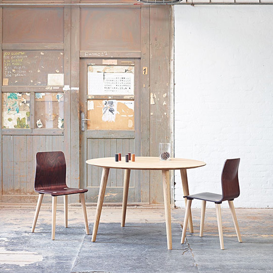 Natural wood table with light finish, paired with two dark brown chairs on a textured floor and a rustic backdrop.