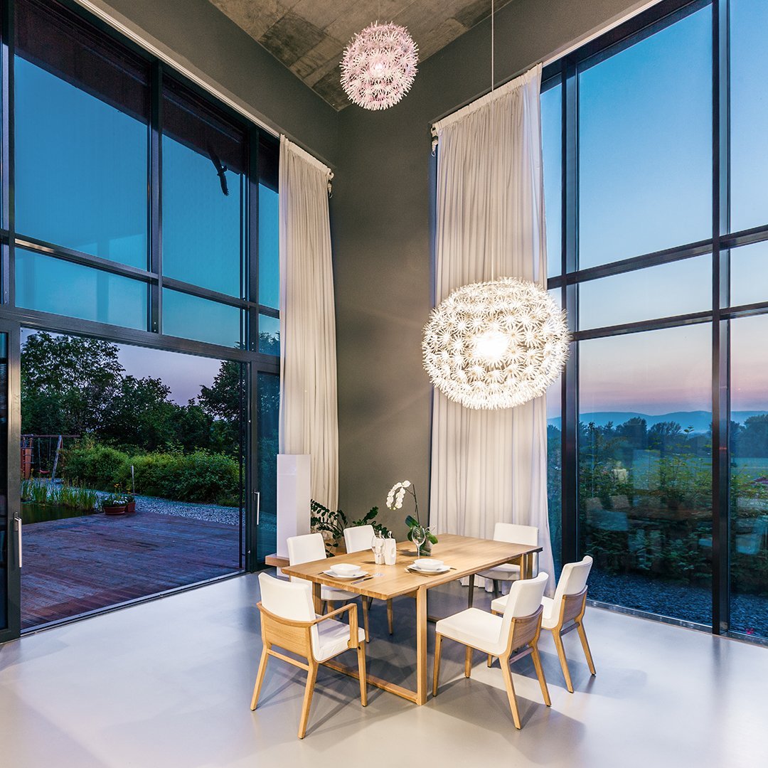 Modern dining area featuring a white, spherical light fixture, wooden table, and minimalist white chairs against large windows.
