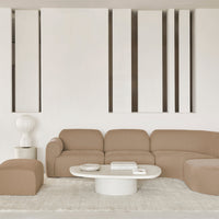 Brown curved sofa and ottoman with a light gray round coffee table on a textured beige rug in a minimalist space.