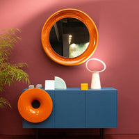 Vibrant orange circular mirror and accents on a dark blue cabinet against a deep burgundy wall with green bamboo.