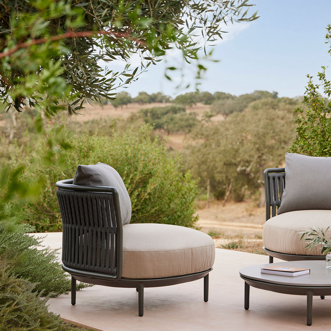 Light gray round outdoor chair with black woven detail and a soft beige cushion, set in a natural green landscape.