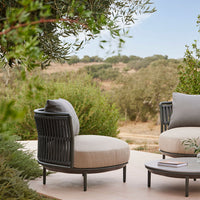 Light gray round outdoor chair with black woven detail and a soft beige cushion, set in a natural green landscape.
