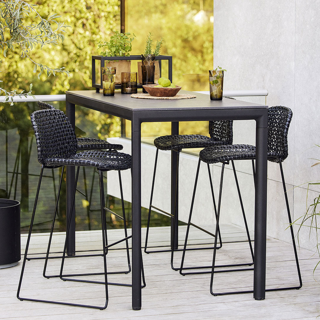 "Stylish black outdoor bar table and chairs with woven patterns, set against a lush green backdrop."
