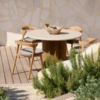 Round beige dining table with natural wood accents, surrounded by light gray cushions and green plants.