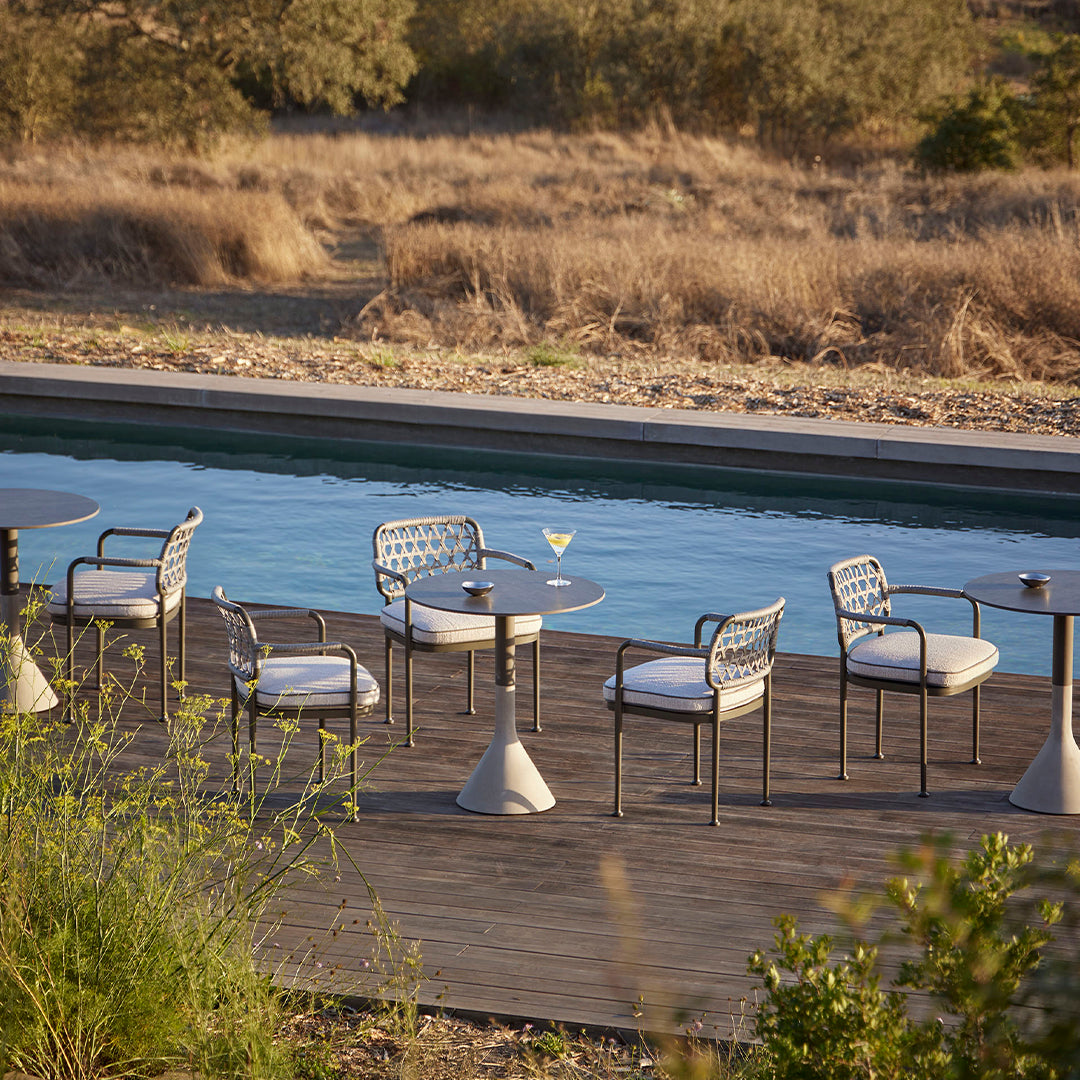 "Outdoor dining set with white cushions and gray metal frames, surrounded by natural earth tones and a serene pool view."