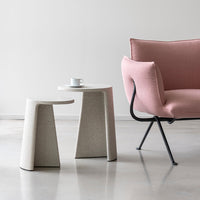 Light pink armchair next to a textured white side table on a sleek gray floor. Modern, minimalistic design.