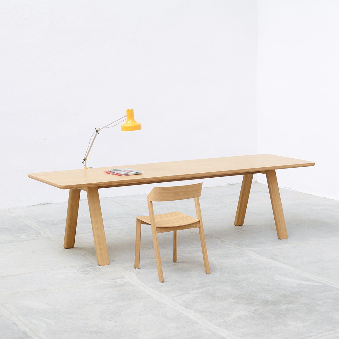 Light wood table and chair set with a bright yellow lamp, minimalist design against a white wall and concrete floor.