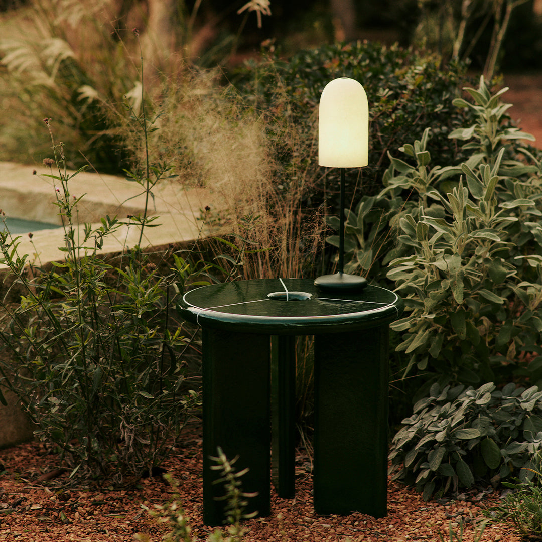 Dark green round side table with white accents, set against a natural garden backdrop of muted greens and soft beige tones.