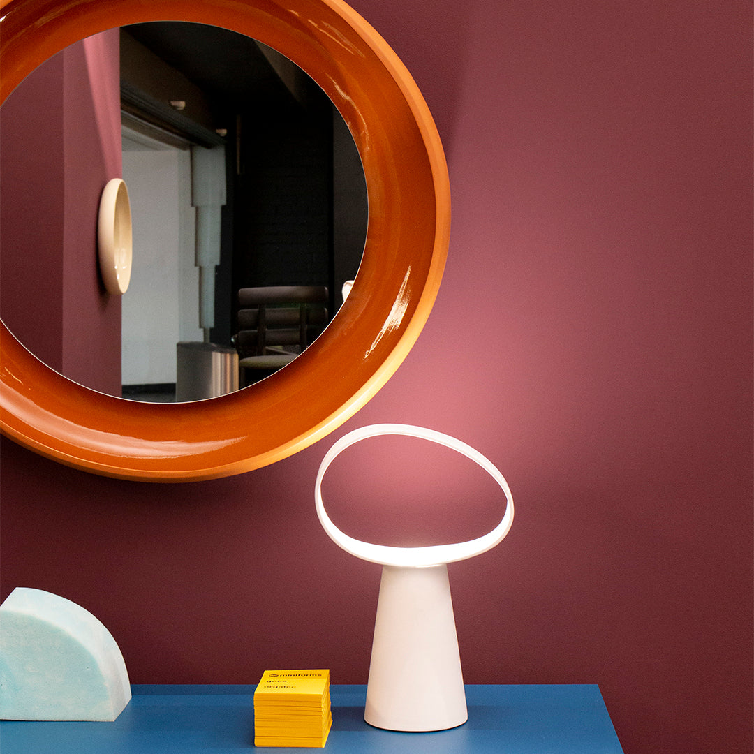 Modern lamp with a white base, set against a deep burgundy wall, featuring an orange circular mirror and blue tabletop.
