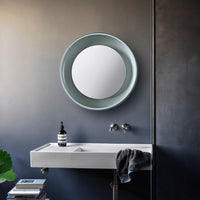 Round, mint green mirror against a dark gray wall, paired with a sleek white sink and a textured charcoal towel.