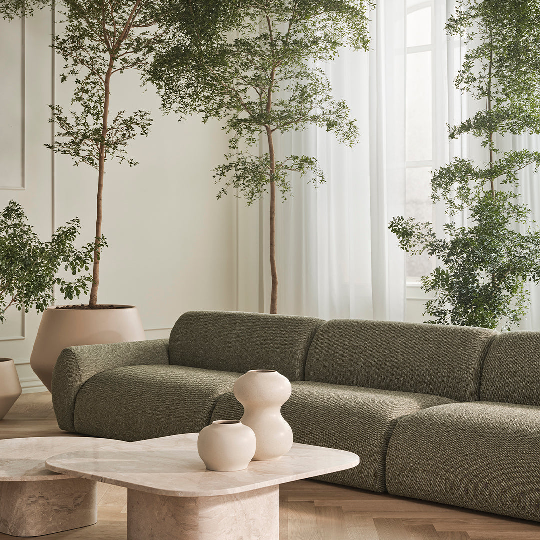 Olive green textured sofa with neutral ceramic vases on a light marble coffee table amid lush greenery.