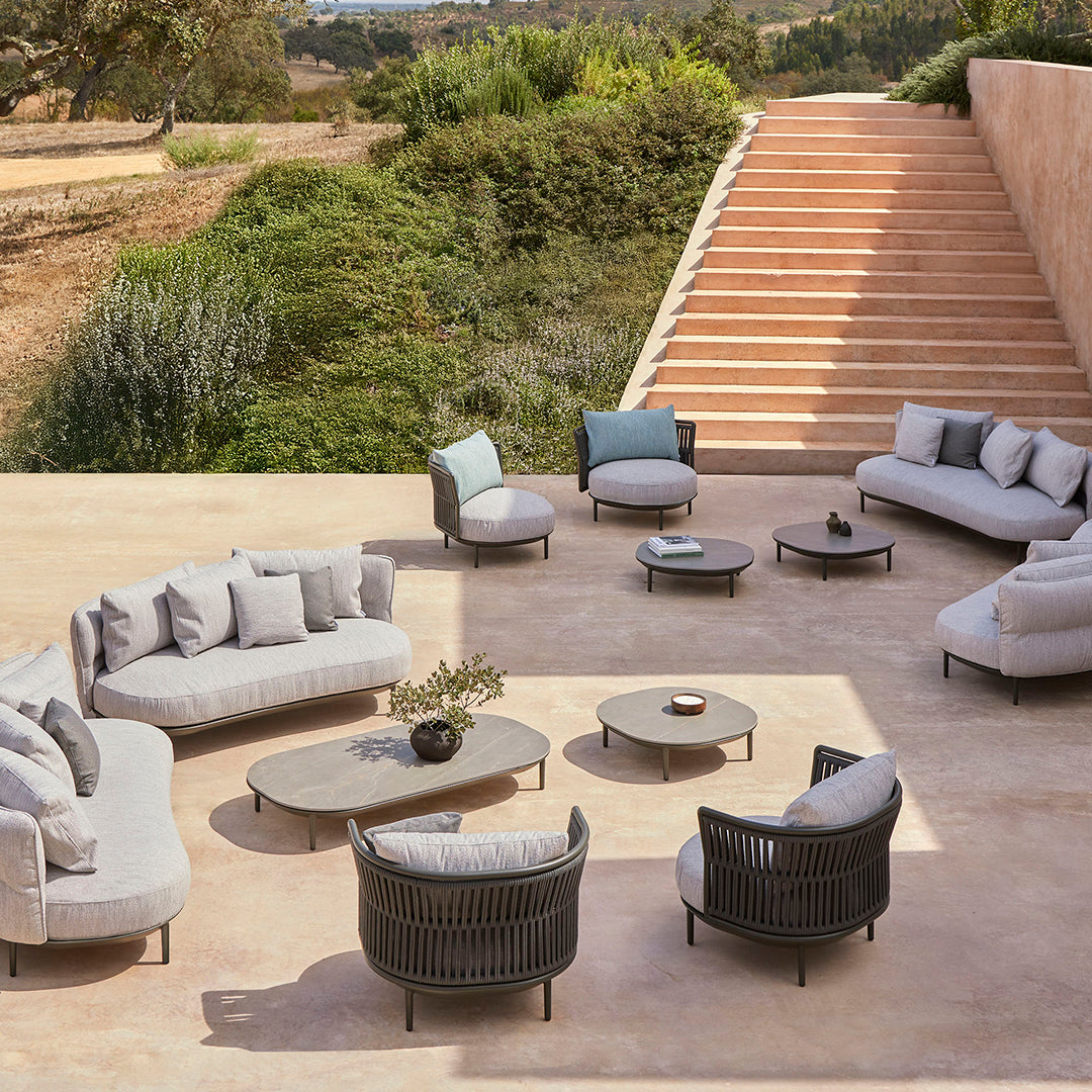 Modern outdoor lounge set with light gray cushions and black wicker accents, arranged on a neutral stone patio.