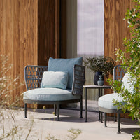 Light blue outdoor chair with textured cushions and black metal accents. Natural wood backdrop and greenery.